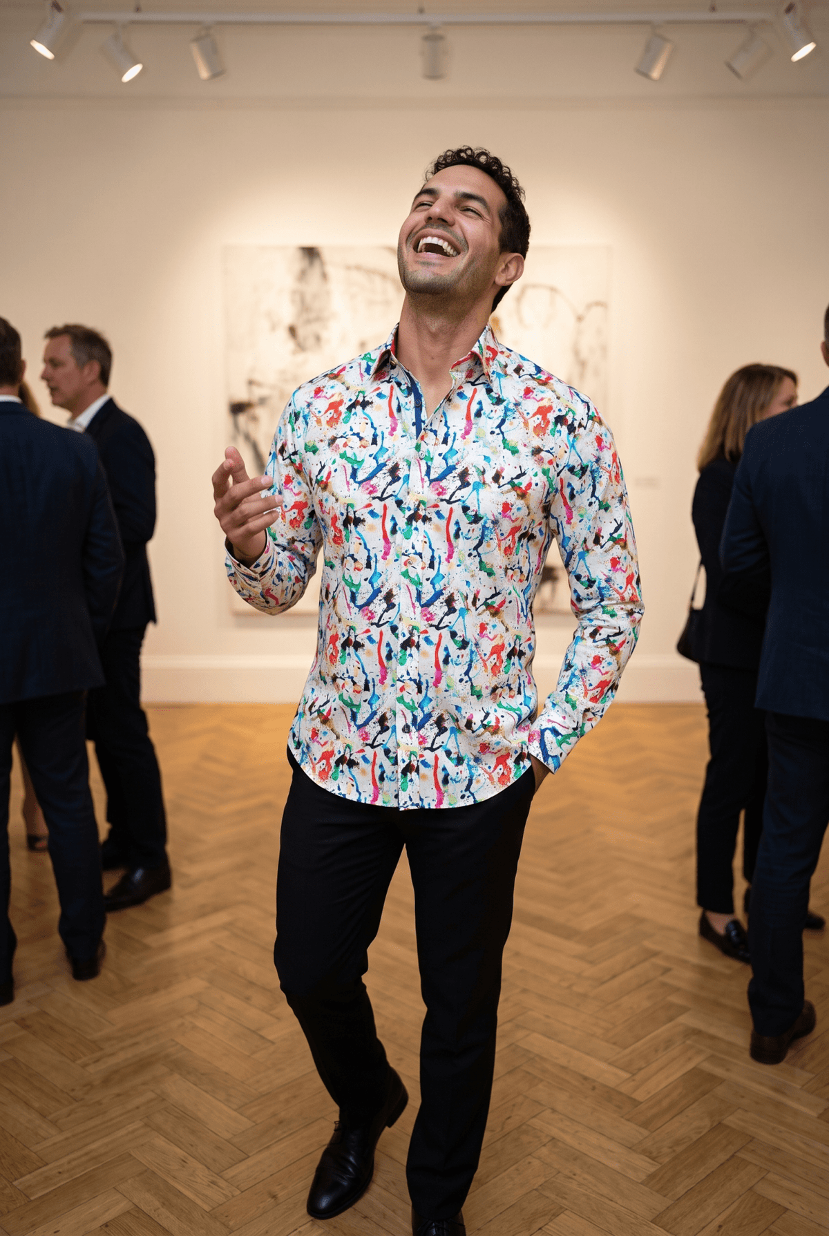 A man in his late twenties wearing the Claudio Lugli Art Attack Paint Splash Print Shirt at a private view in a tall Georgian gallery, head tipped back mid laugh with one hand raised, parquet floor and blurred guests behind