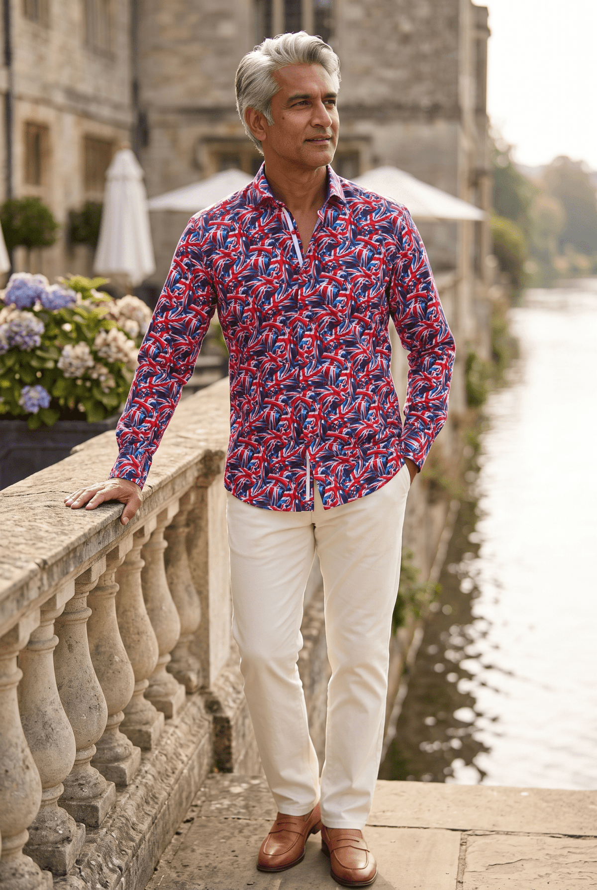 A man in his fifties wearing the Claudio Lugli Union Jack Print Shirt on a riverside members club terrace on a hazy summer afternoon, standing side on at the balustrade with one hand in his pocket, parasols and hydrangeas behind him