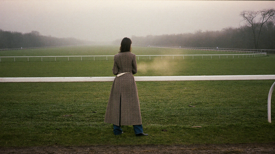 A woman in a long herringbone tweed riding jacket at a misty Gloucestershire racecourse in November - the winter racing wardrobe
