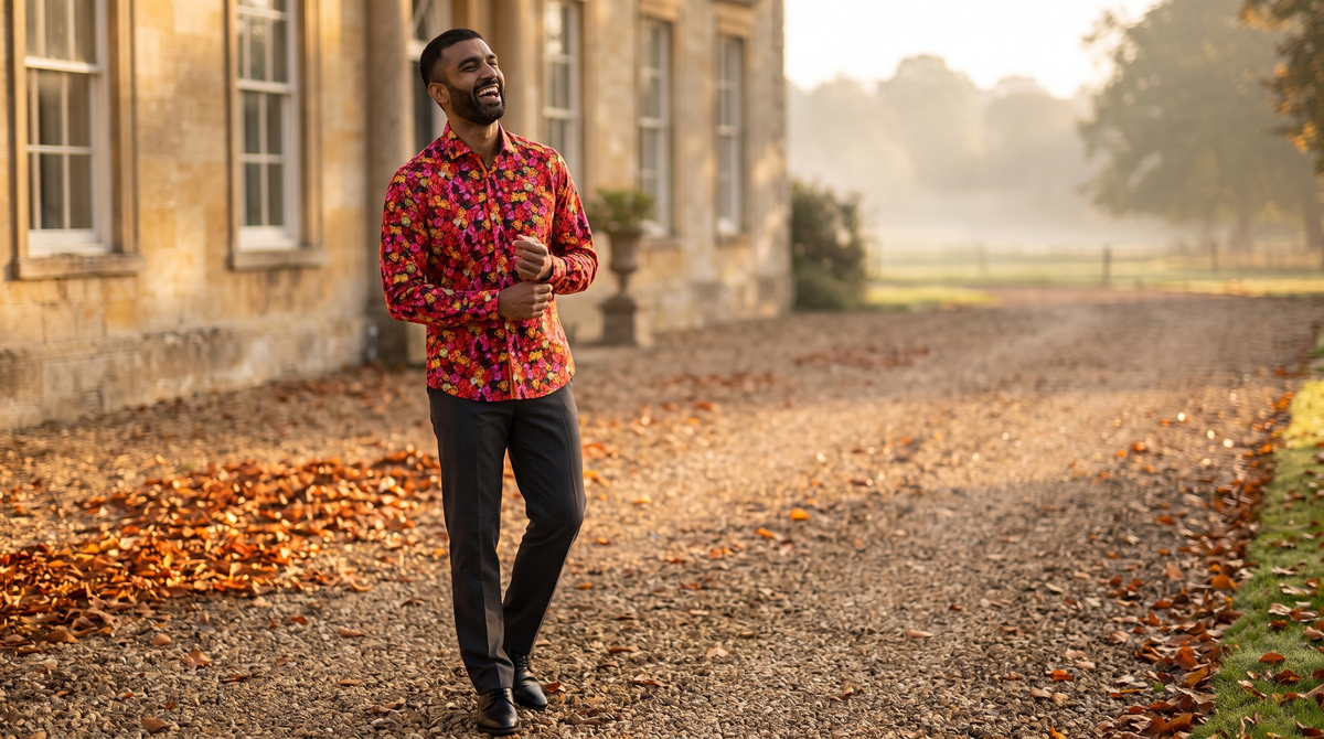 Autumn wedding guest outfit men: South Asian man late thirties, cropped beard, head tipped back mid-laugh adjusting a cuff on a country-house gravel drive at golden hour, Claudio Lugli Midnight Rose print shirt buttoned to the collar, charcoal trousers