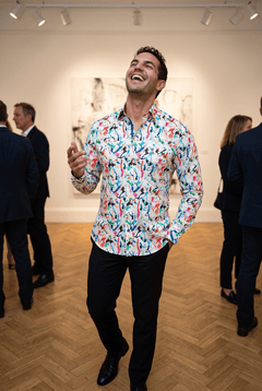 A man in his late twenties wearing the Claudio Lugli Art Attack Paint Splash Print Shirt at a private view in a tall Georgian gallery, head tipped back mid laugh with one hand raised, parquet floor and blurred guests behind