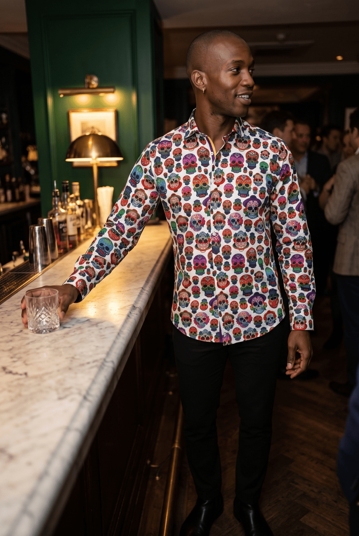 A man in his early thirties wearing the Claudio Lugli Embroidery Skulls Shirt in a dark panelled Soho members bar late at night, setting a glass down on marble with one arm still extended, brass picture lamp and deep green walls behind