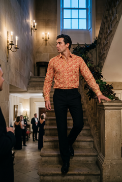 A man in his late twenties wearing a Claudio Lugli Baroque Legacy Collection print shirt at a black tie evening, coming down the stone staircase of Georgian assembly rooms past candle sconces, the cream ground and scattered red motifs reading clearly