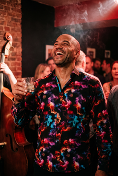 A man in his early forties wearing the Claudio Lugli Smoke Print Shirt in Multicolour at a Soho basement jazz club at midnight, glass raised and head tipped back laughing, vivid magenta cyan and orange smoke reading against the black ground under the stage wash