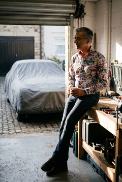 A man in his early fifties wearing the Claudio Lugli Monaco Grand Prix Print Shirt in a mews garage on a cold autumn morning, sitting on the bench edge with a mug in both hands, the pale ground and small rectangular vintage race poster panels reading across the shirt