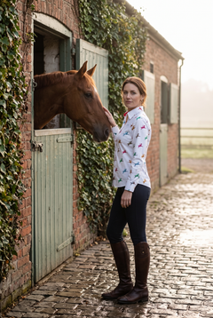 A woman in her late thirties wearing the Claudio Lugli Horse and Rider Womens Print Shirt in a misty stable yard on an October morning, one hand on a chestnut horse's neck and looking back over her shoulder, small multicoloured horse and rider motifs on a white ground with a red inner collar