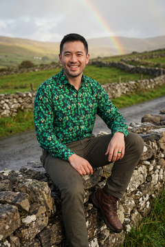 Four Leaf Clover Emerald Print Shirt by Claudio Lugli – St Patrick's Day outfit idea for men. Sitting on a dry-stone wall under a rainbow, west of Ireland. Model: Irish-Chinese man, 33, short dark hair, stubble, medium build. Designed in London, sizes S to 6XL.