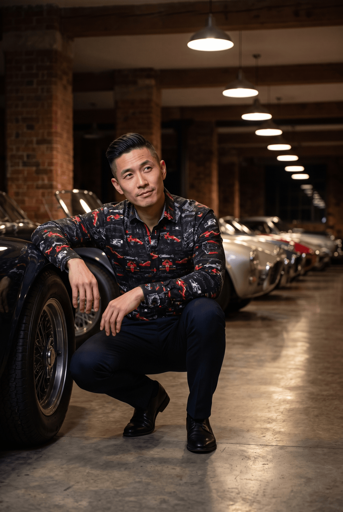 A man in his late thirties wearing the Claudio Lugli Pit Lane Proto-Type F1 Print Shirt, crouched low beside the front wheel of a classic racing car in a converted warehouse at night, looking up past the camera under pendant lights