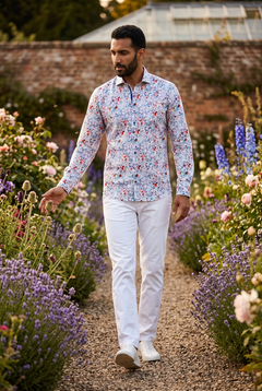A man in his early thirties wearing the Claudio Lugli Wild Bloom Print Shirt, mid stride on a gravel path in a walled English garden in high summer, brushing a flower head with one hand, glasshouse beyond