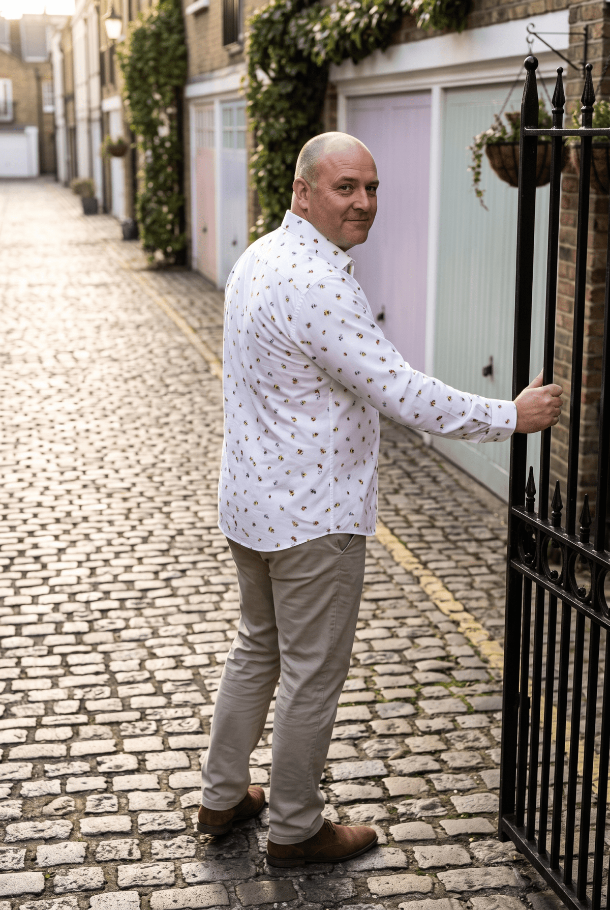 A man in his late forties wearing the Claudio Lugli Worker Bee Print Shirt in a cobbled Mayfair mews on a bright morning, turning back over one shoulder with a hand on the gate, painted garage doors and low sun down the mews