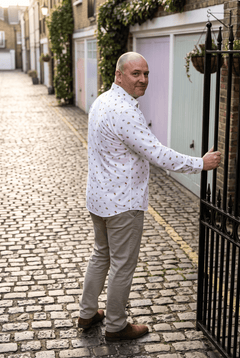 A man in his late forties wearing the Claudio Lugli Worker Bee Print Shirt in a cobbled Mayfair mews on a bright morning, turning back over one shoulder with a hand on the gate, painted garage doors and low sun down the mews